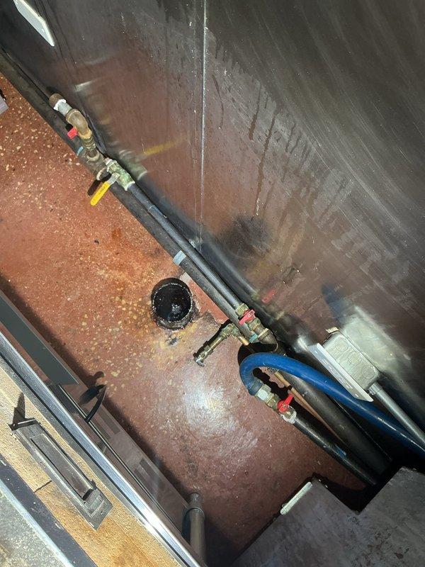 Responded to customer request for hydrojetting estimate after experiencing water backups from floor drains when operating certain sinks. Inspected commercial kitchen area including stainless steel sink units, visible floor drains, and adjacent plumbing infrastructure. Identified grease trap requiring maintenance and documented evidence of water damage and oxidation in the utility area where pipe assemblies connect to drainage system.