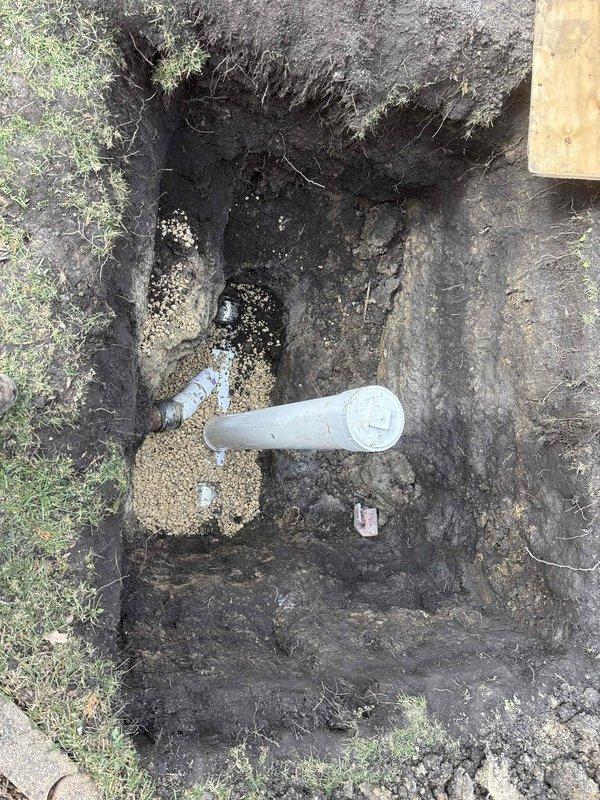 Excavated access pit to expose water/drainage line where significant corrosion was identified on copper T-junction fitting. Documented standing water accumulation at excavation site, likely indicating leak from deteriorated pipe section. Replacement parts and financing were arranged to resolve the drainage issue.