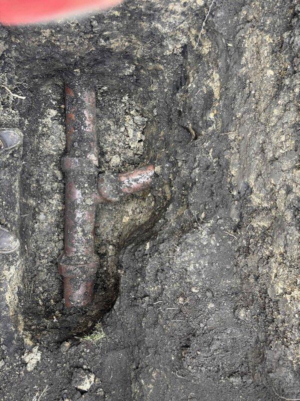 Excavated access pit to expose water/drainage line where significant corrosion was identified on copper T-junction fitting. Documented standing water accumulation at excavation site, likely indicating leak from deteriorated pipe section. Replacement parts and financing were arranged to resolve the drainage issue.
