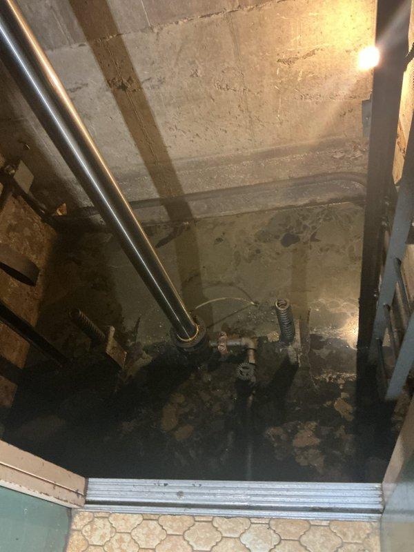 Conducted site assessment for sump pump installation in utility area with recurring water backup issues. Observed standing water on concrete floor with exposed plumbing components and vertical metal pipes along concrete block walls. Documented evidence of frequent flooding in the mechanical space, confirming customer's report of pit that backs up regularly and requires proper drainage solution.