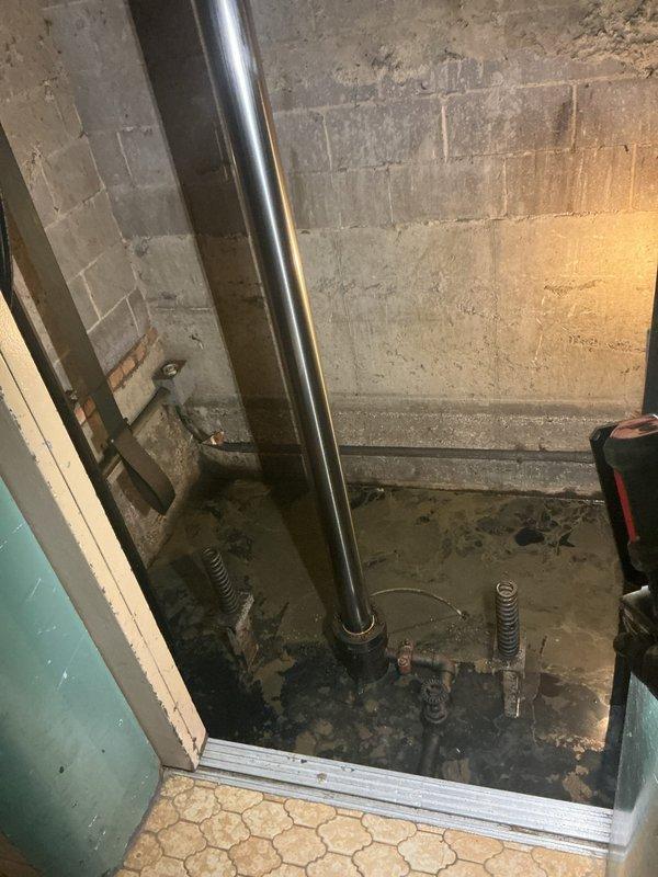 Conducted site assessment for sump pump installation in utility area with recurring water backup issues. Observed standing water on concrete floor with exposed plumbing components and vertical metal pipes along concrete block walls. Documented evidence of frequent flooding in the mechanical space, confirming customer's report of pit that backs up regularly and requires proper drainage solution.