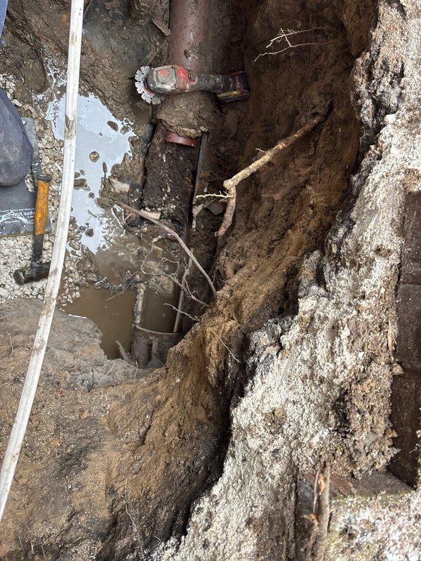 Conducted camera inspection of underground pipe system in excavated trench, documenting significant corrosion and deterioration of metal piping. Identified standing water accumulation at pipe connection point, likely indicating leak or drainage issue requiring repair. Visual examination revealed valve connection point with extensive rust damage requiring replacement.