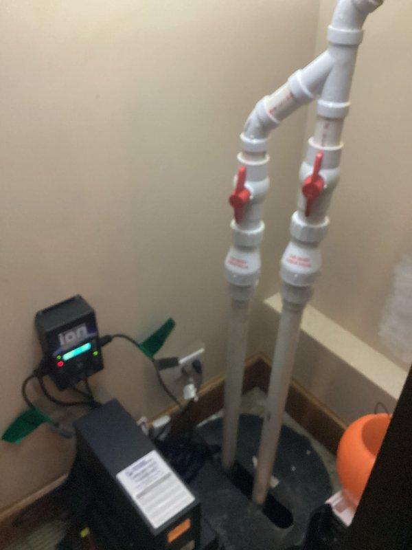 Installed Liberty PC backup sump pump system in basement, including dual PVC discharge lines with check valves and a wall-mounted electronic control unit with digital monitoring display. System features battery backup capability and was fully tested to ensure proper operation during power outages.