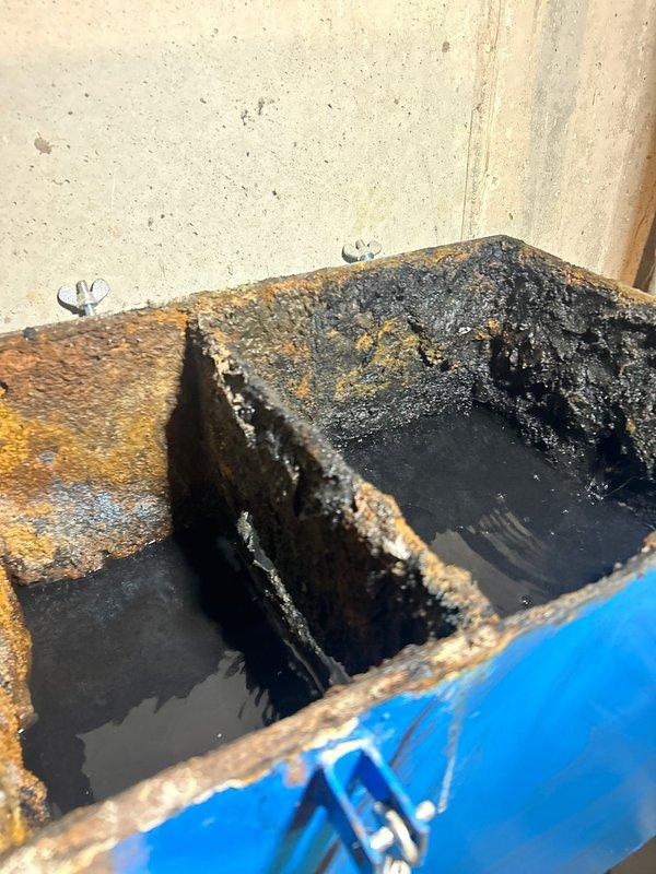 Responded to service call regarding clogged grease trap in 35-year-old home basement. Upon inspection, discovered severely corroded catch basin with significant rust deterioration, standing water, and compromised metal dividers. Cleared blockage, documented extensive deterioration with photos showing the damaged internal components and external housing, and recommended replacement due to advanced corrosion affecting system functionality.