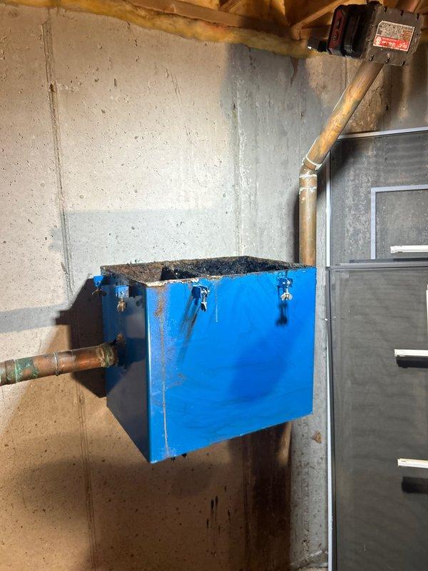 Responded to service call regarding clogged grease trap in 35-year-old home basement. Upon inspection, discovered severely corroded catch basin with significant rust deterioration, standing water, and compromised metal dividers. Cleared blockage, documented extensive deterioration with photos showing the damaged internal components and external housing, and recommended replacement due to advanced corrosion affecting system functionality.