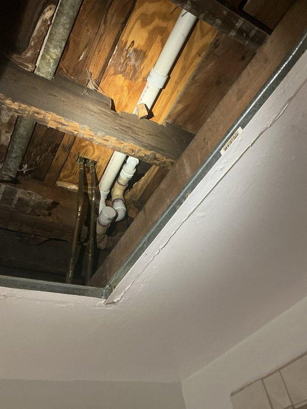 Accessed ceiling cavity to inspect plumbing system, revealing both PVC and copper piping installations. Observed water staining on wooden joists and potential corrosion on copper pipes, indicating previous moisture exposure in the structural framework. Completed installation of new PVC drain line system with appropriate elbows and connections through the ceiling joists.