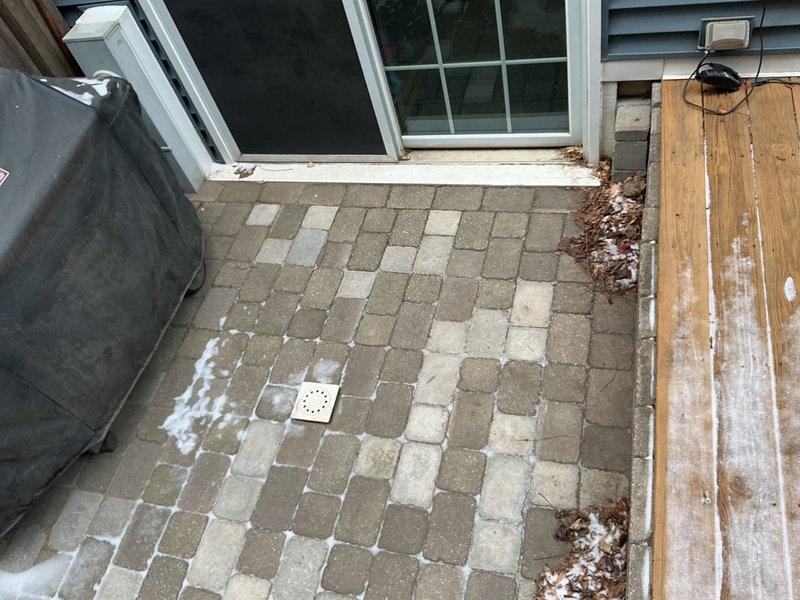 Conducted inspection of outdoor patio drainage system where water was slowly seeping through cracked drain. Documented square drainage cover embedded in stone pavers with visible deterioration around edges. No active backup was observed at time of assessment.