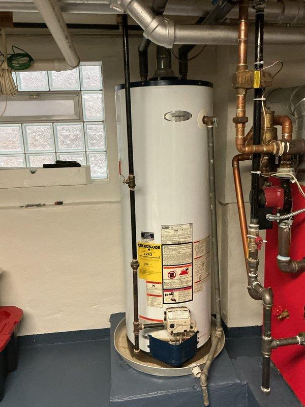 Conducted a comprehensive estimate for replacing an aging Whirlpool water heater installed in the utility room. Inspected the existing plumbing configuration, which includes a mix of galvanized steel, copper, and black pipes with multiple connection points and a prominent blue valve handle. Noted the current unit sits on a metal catch pan and would require coordination with the adjacent heating system components when performing the replacement.