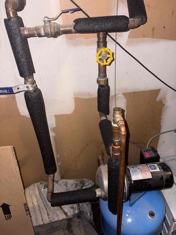 Responded to service call for low water pressure and pipe banging issues. Inspected plumbing system including insulated vertical pipes, copper lines, and pressure tank with attached pump. Diagnosed and adjusted the yellow flow control valve to restore proper water pressure and eliminate water hammer effect causing the sporadic banging noises.