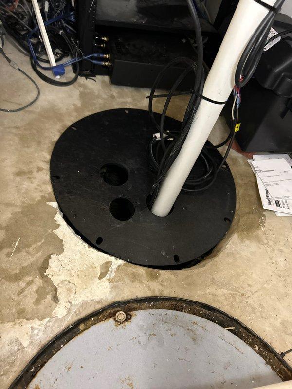 Conducted site assessment of basement sump pump system, documenting existing installation with black circular base plate showing corrosion and moisture issues. Evaluated current PVC discharge pipe configuration, noting S-shaped routing through floor joists with multiple elbow joints and connection points requiring attention. Scheduling immediate follow-up pending B-box discovery to implement proper waste pump solution.