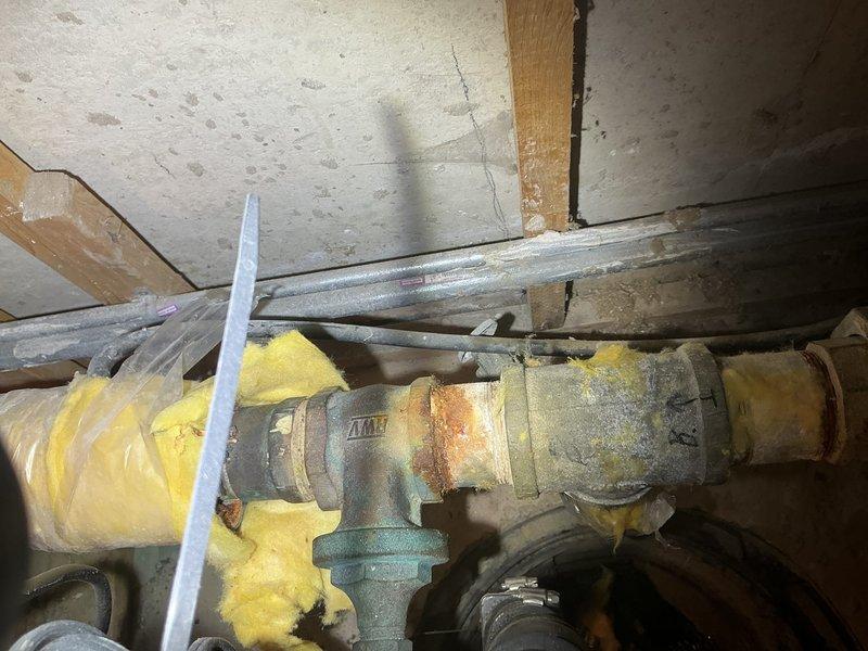 Conducted inspection of customer's water meter piping system. Identified significant corrosion and deterioration on metal pipe junction with T-shaped valve, showing rust and greenish-blue patina. Documented existing Quiet Check Valve installation and noted damaged yellow fiberglass insulation surrounding portions of the piping system. Provided comprehensive estimate for replacement of corroded components to prevent potential future leaks.