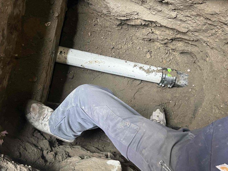 Completed installation of sewer line by excavating a trench and installing NAPCO PVC pipe with proper metal coupling. Connected white PVC pipe to existing copper line using appropriate metal fitting with green indicator tag, ensuring proper alignment and secure connections for optimal drainage performance.