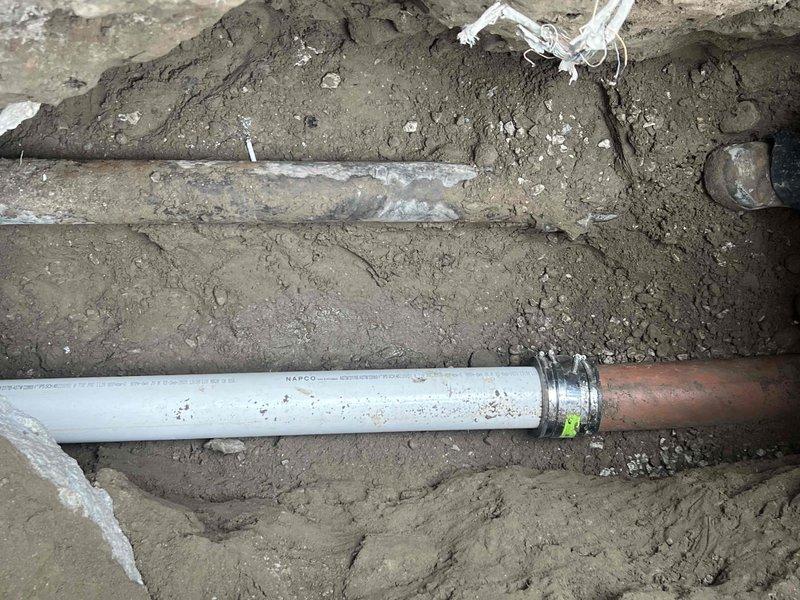 Completed installation of sewer line by excavating a trench and installing NAPCO PVC pipe with proper metal coupling. Connected white PVC pipe to existing copper line using appropriate metal fitting with green indicator tag, ensuring proper alignment and secure connections for optimal drainage performance.