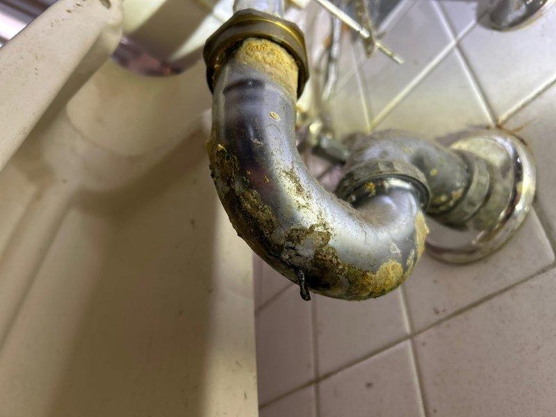 Located and identified source of bathroom sink leak as severely deteriorated drain pipe with significant corrosion and mineral buildup. Documented active water leakage from the bottom of the P-trap assembly, which showed multiple areas of deterioration including yellowish-brown deposits at the fixture connection point and extensive oxidation throughout the curved section.