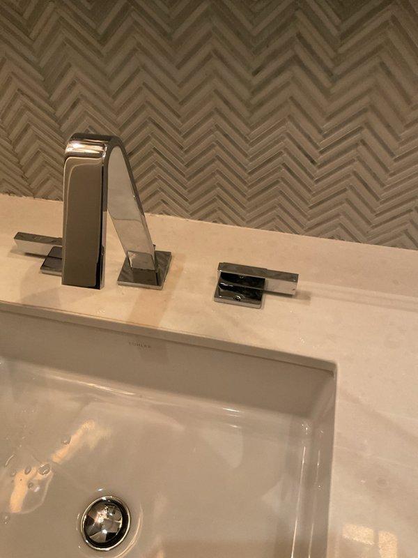 Successfully replaced broken bathroom faucet with a new chrome model featuring angular design and separate handle components, installing it properly on the light beige countertop with the decorative herringbone backsplash. Also inspected and repaired kitchen faucet drip issue located above stove.