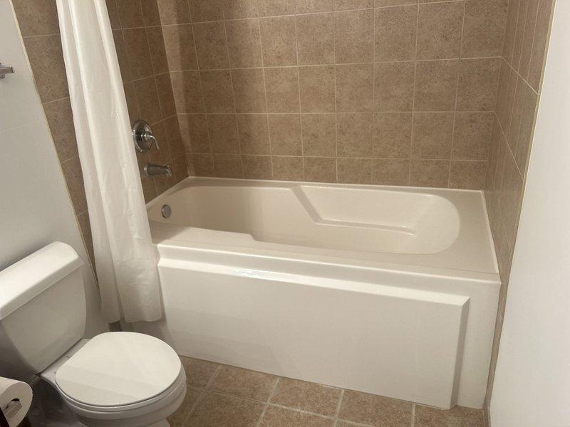 Performed bathroom remodel estimate for residential client. Inspected existing bathroom features including white porcelain bathtub with chrome fixtures, white toilet, and beige/tan tile surround extending from floor to walls. Documented current condition of fixtures and tile work with photos for estimate preparation.