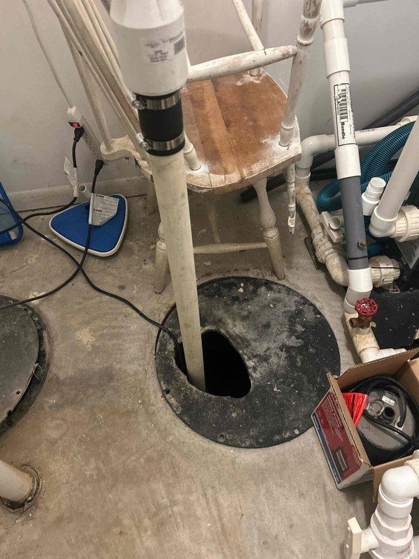 Completed inspection of basement sump pump installation with associated PVC drainage system. Documented condition of shutoff valve and pipe connections including the black insulated vertical pipes with red valve component. Verified proper installation of flexible drainage hose and all plumbing connections, with no visible leaks or deterioration observed in the utility area infrastructure.