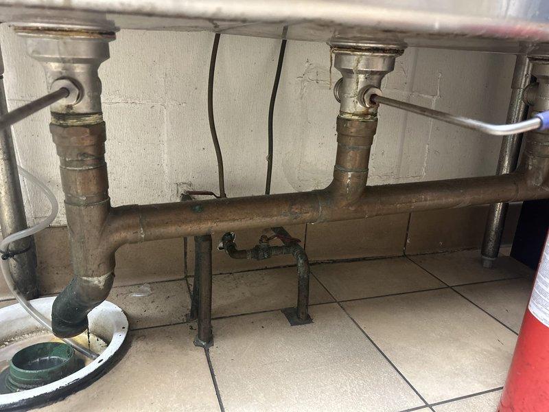 Responded to service call for commercial grease trap cleaning. Inspected plumbing infrastructure including copper piping system with visible oxidation at joints and connection points. Serviced floor drain showing accumulated sediment and standing water, cleaning the green threaded fitting and ensuring proper drainage functionality.