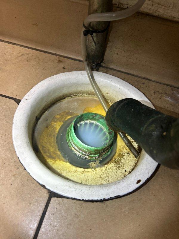 Responded to service call for commercial grease trap cleaning. Inspected plumbing infrastructure including copper piping system with visible oxidation at joints and connection points. Serviced floor drain showing accumulated sediment and standing water, cleaning the green threaded fitting and ensuring proper drainage functionality.