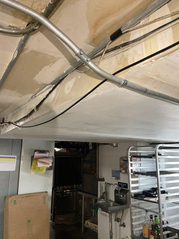 Responded to service call regarding floor drain backup and two problematic sinks. Upon inspection, discovered significant water damage around a wall access panel where a copper pipe with a green shut-off valve was visible. Further examination of the commercial kitchen area revealed exposed metal conduits and electrical wiring running along the ceiling, with evidence of unfinished drywall work suggesting previous plumbing issues. Cleared the floor drain obstruction and serviced both sink drains to restore proper drainage function.