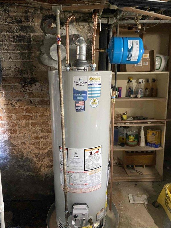 Completed inspection of an aging Bradford White water heater installed against brick wall with copper piping showing patina development. Observed existing installation including metallic vent pipe with elbow joint, brass valve connections, and proper ventilation components. Provided financing options for replacement of this unit which appears to be from 2012 based on visible date marking.