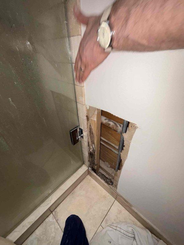 Performed inspection of suspected water leak behind shower enclosure. Removed drywall section to access the area, revealing evidence of moisture damage to wooden framing members. Documented extent of deterioration and confirmed active water intrusion in the shower corner where tiled wall meets painted drywall, likely originating from compromised waterproofing at the metal edge trim junction.