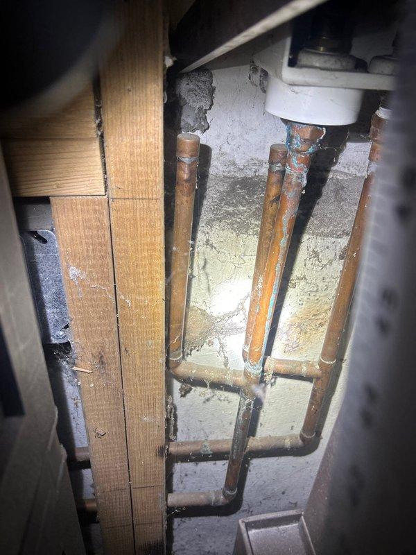Conducted inspection of residential drainage system, identifying significant copper pipe corrosion and mineral buildup in utility area. Observed compromised pipe integrity with greenish-blue patina formation and deterioration at connection points. Basement area shows evidence of moisture issues with debris accumulation and potential water damage to surrounding concrete surfaces.