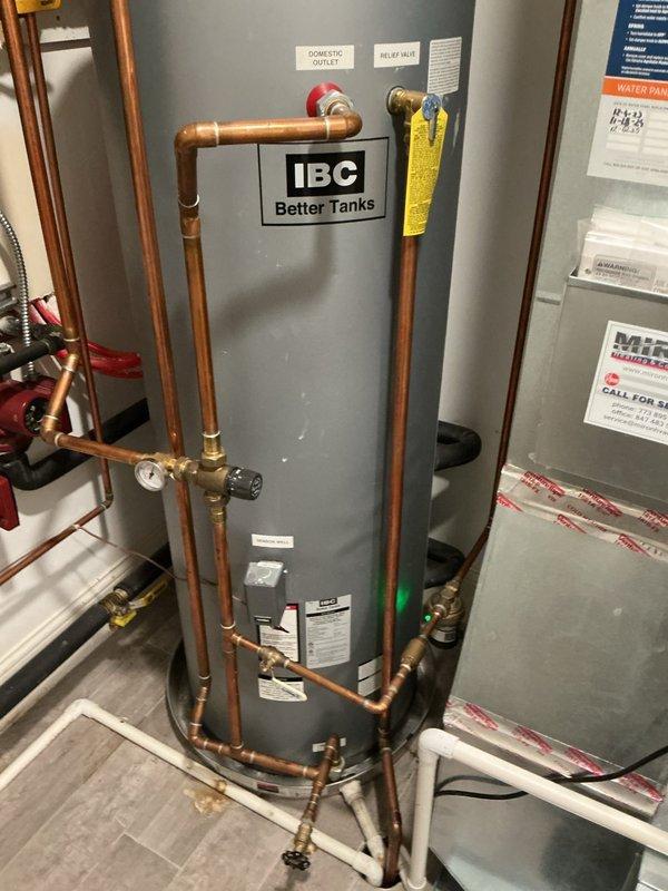 Successfully installed an IBC "Better Tanks" water heater in utility area, complete with proper copper piping connections, pressure gauge, and relief valve. System features domestic outlet connections and appropriate drainage via PVC piping, all mounted securely on a protective floor pan with all manufacturer labels and information tags properly affixed.