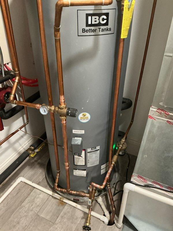 Successfully installed an IBC "Better Tanks" water heater in utility area, complete with proper copper piping connections, pressure gauge, and relief valve. System features domestic outlet connections and appropriate drainage via PVC piping, all mounted securely on a protective floor pan with all manufacturer labels and information tags properly affixed.