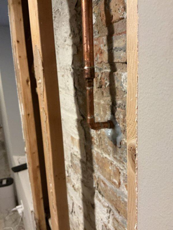 Completed installation of a new copper supply line with brass fittings and shutoff valve along exposed brick wall. Existing drywall was removed to access the plumbing system, revealing deteriorated mortar around the brick structure. New blue-handled shutoff valve was successfully installed and tested for proper operation.