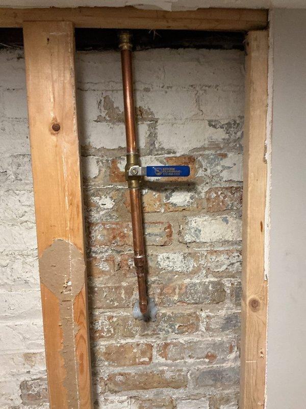 Completed installation of a new copper supply line with brass fittings and shutoff valve along exposed brick wall. Existing drywall was removed to access the plumbing system, revealing deteriorated mortar around the brick structure. New blue-handled shutoff valve was successfully installed and tested for proper operation.