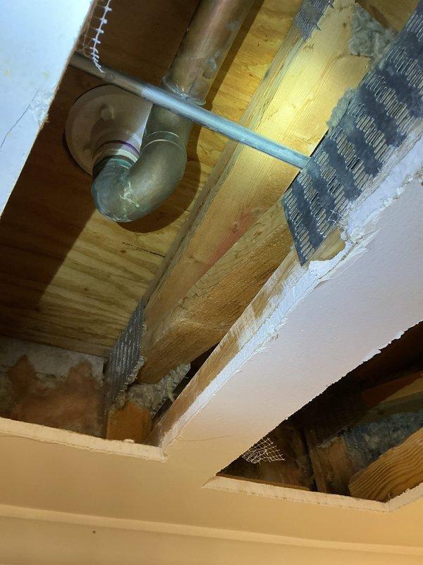 Opened ceiling beneath shower to inspect plumbing issues, revealing water damage to wooden joists and a corroded copper pipe with temporary tape repair. Identified leaking P-trap connection to the main drain line and evidence of previous moisture infiltration requiring replacement of affected plumbing components.