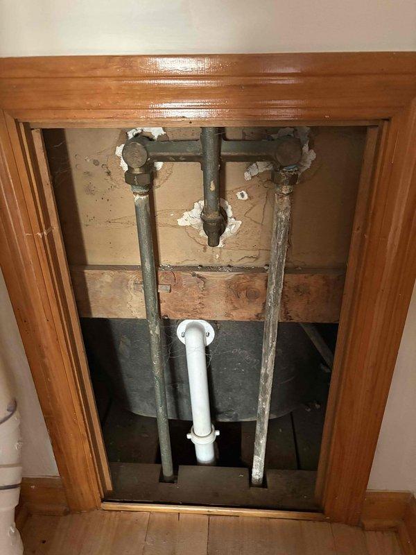 Inspected exposed plumbing system in wall access panel, documenting significant corrosion on galvanized steel pipes alongside newer PVC installation, with notable wall damage around pipe penetrations. Evaluated bathroom sink fixture with chrome faucet mounted on teal tile wall to provide comprehensive estimate for recommended repair and replacement options.