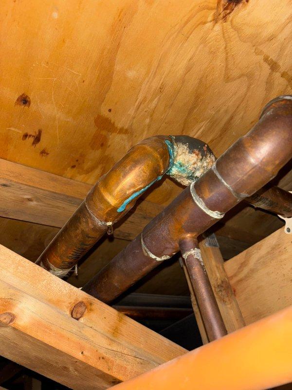 Located and identified source of basement ceiling leak as a corroded copper pipe fitting. Inspection revealed significant verdigris (blue-green oxidation) and moisture accumulation at a 90-degree elbow joint in the copper water supply line, with visible water staining on surrounding wooden framing members indicating ongoing leakage. Water remains shut off to prevent further damage.