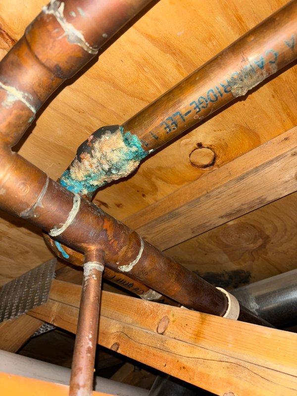 Located and identified source of basement ceiling leak as a corroded copper pipe fitting. Inspection revealed significant verdigris (blue-green oxidation) and moisture accumulation at a 90-degree elbow joint in the copper water supply line, with visible water staining on surrounding wooden framing members indicating ongoing leakage. Water remains shut off to prevent further damage.