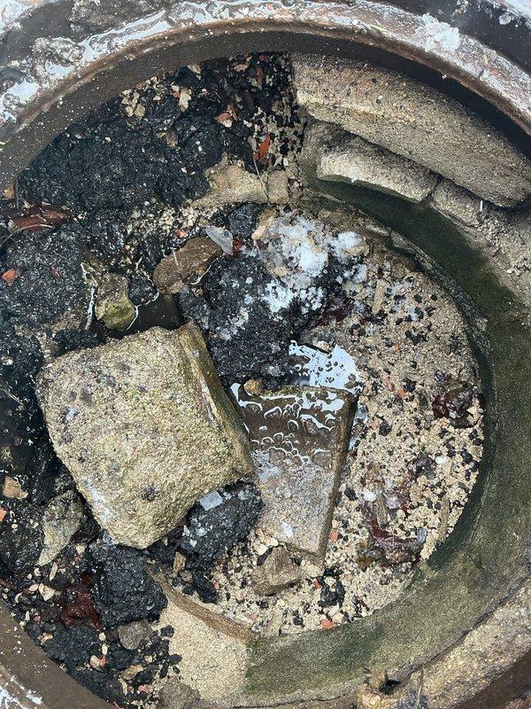 Responded to service call for manhole filled with debris. Upon inspection, discovered significant accumulation of construction waste including concrete blocks, stone fragments, and sand causing complete blockage. Standing water was present with visible algae growth on interior walls indicating prolonged moisture exposure and drainage issues.