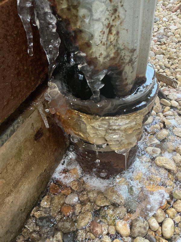 Conducted inspection of exterior drainage system, identifying two severely compromised downspouts requiring replacement. First downspout exhibited extensive ice formation and corrosion at pipe joints with visible water pooling and freezing at the base, while the second downspout showed significant aging with corrosion, greenish algae growth, and deterioration at the ground connection point. Recommended complete replacement of both downspouts to prevent structural water damage and ensure proper drainage.
