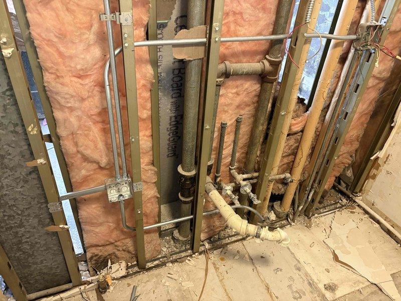 Inspected existing plumbing infrastructure for kitchen and bathroom remodel project. Documented exposed wall cavities showing R-11 insulation, horizontal white supply lines with sealant at connection points, and vertical drain stack with multiple PVC and copper pipes running through wooden framing. Assessment completed to determine requirements for upcoming plumbing modifications.