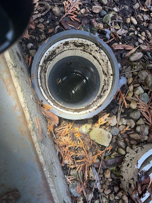 Inspected exterior drain tile system showing significant buildup and staining in the pipe interior. Observed standing water at the bottom of the drainage pipe and documented condition of the dual drainage system consisting of a black circular pipe and white slotted drain cover, both surrounded by pebbles and organic debris accumulation.