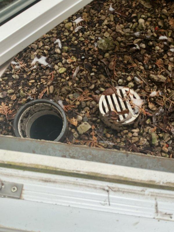 Inspected exterior drain tile system showing significant buildup and staining in the pipe interior. Observed standing water at the bottom of the drainage pipe and documented condition of the dual drainage system consisting of a black circular pipe and white slotted drain cover, both surrounded by pebbles and organic debris accumulation.