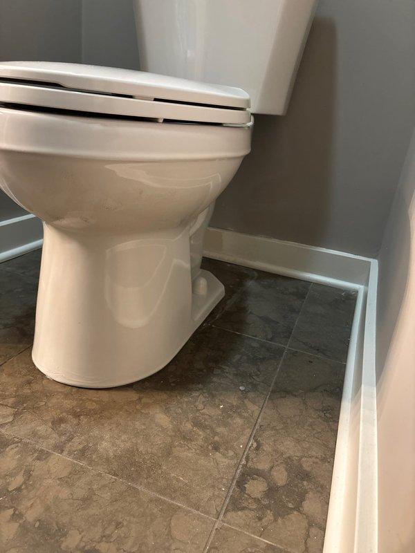 Successfully completed toilet installation in bathroom with light gray walls and brown tile flooring. Installed new white porcelain toilet with standard design, connecting braided stainless steel supply line to existing chrome shutoff valve. Noted minor water staining around toilet base that was present prior to installation.