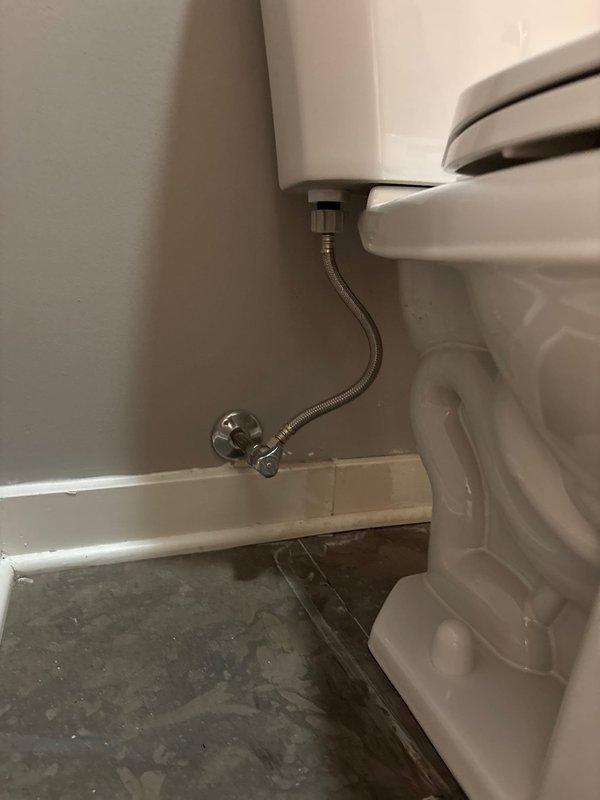 Successfully completed toilet installation in bathroom with light gray walls and brown tile flooring. Installed new white porcelain toilet with standard design, connecting braided stainless steel supply line to existing chrome shutoff valve. Noted minor water staining around toilet base that was present prior to installation.