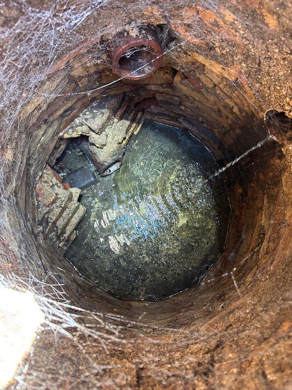 Performed inspection of 100-year-old main drain line with catch basin, documenting significant deterioration including brick wall degradation, standing water accumulation, and mineral deposits. Observed basement area showing signs of moisture intrusion with possible seepage through foundation walls, requiring further evaluation for hydro jetting service as requested.