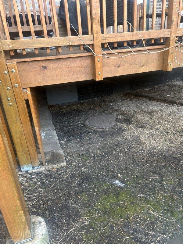 Performed on-site assessment for hydrojetting service request. Inspected outdoor drainage system, including circular utility drain cap embedded in ground beneath wooden deck structure. Examined concrete floor with visible cracks showing signs of water damage and identified potential access point for service.