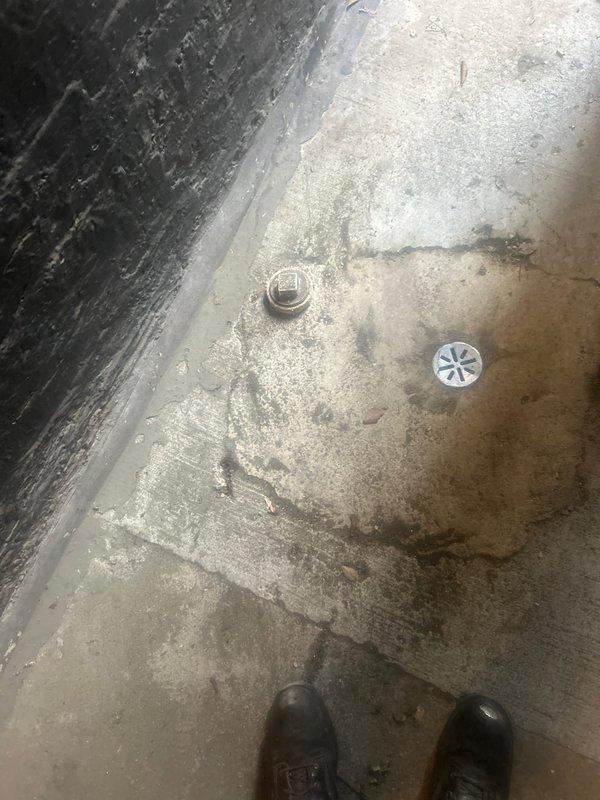 Performed on-site assessment for hydrojetting service request. Inspected outdoor drainage system, including circular utility drain cap embedded in ground beneath wooden deck structure. Examined concrete floor with visible cracks showing signs of water damage and identified potential access point for service.