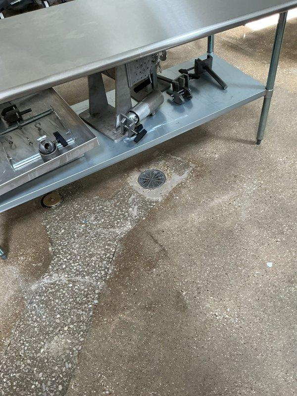 Inspected commercial facility experiencing fly infestation from floor drain. Documented corroded plumbing infrastructure, specifically a deteriorating valve assembly with significant rust buildup at connection points. Observed standing water around floor drain in food preparation area, which likely serves as breeding ground for insects.