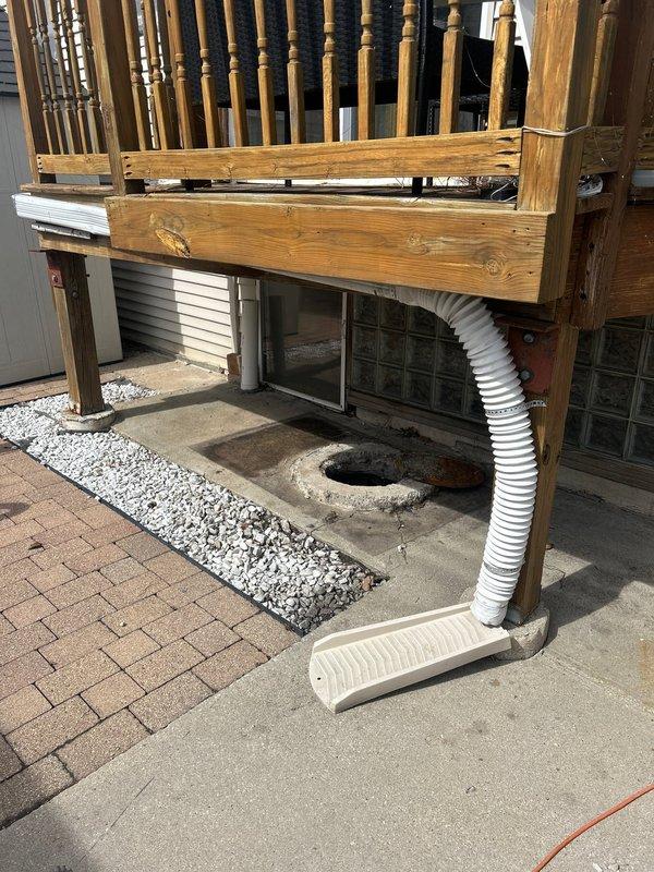Conducted thorough inspection and provided estimate for catch basin cleaning service. Examined drainage system including deck area with circular concrete opening and connected white flexible duct/hose. Assessed associated components including downspout connection and surrounding drainage infrastructure under wooden deck structure.