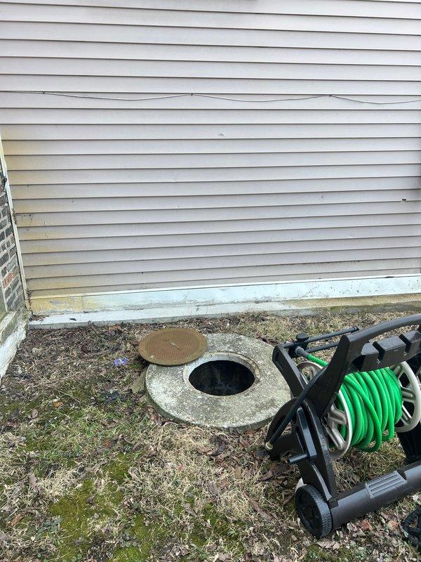 Performed inspection for catch basin cleaning service in customer's absence. Accessed the circular concrete catch basin with metal cover located adjacent to the residence's foundation. Documented current condition with photographs showing the basin area, surrounding property features, and access points for future service planning.