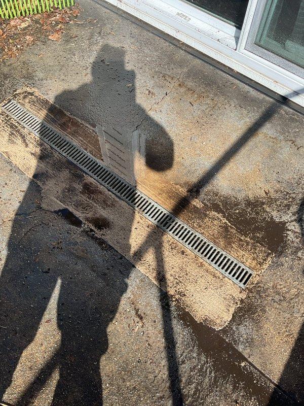 # Job Description

Responded to service call for backed-up exterior drain system. Upon arrival, located linear trench drain with metal grate installed in concrete patio area adjacent to sliding glass door entry. Drain showed visible signs of water backup and debris accumulation around grate openings.

Performed rodding service on exterior drainage line to clear obstruction causing backup. Inserted drain cable through grate openings and worked through blockage in underground drainage pipe. Removed accumulated organic debris, sediment, and buildup restricting water flow. Verified proper drainage function by testing water flow through system after clearing obstruction.

Inspected drainage grate and surrounding concrete surface for damage. Noted weathered concrete conditions and typical organic debris accumulation but found no structural issues requiring immediate attention. Grate remained securely in place with proper alignment for continued function.

Service restored normal drainage ope