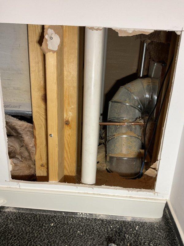 Responded to report of ceiling leak with bathroom located directly above affected area. Upon inspection, accessed vanity cabinet and wall cavity to investigate source of water intrusion. Removed section of drywall to expose plumbing lines, framing, and HVAC ductwork. Identified leak origin in plumbing system above ceiling level. Inspected PVC drain lines and supply piping for signs of failure, corrosion, or loose connections. Documented conditions with photographs showing cabinet interior, exposed plumbing penetrations, and compromised wall cavity with visible moisture damage to ductwork and surrounding materials.
